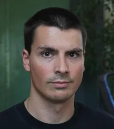 Friedrich Gies im Portrait, in schwarzem T-Shirt vor einer grünen Metalltür.