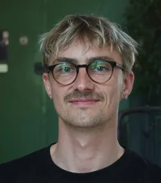 Lucas Pilzen im Portrait, mit Brille und in schwarzem T-Shirt vor einem dunkelgrünen Hintergrund.